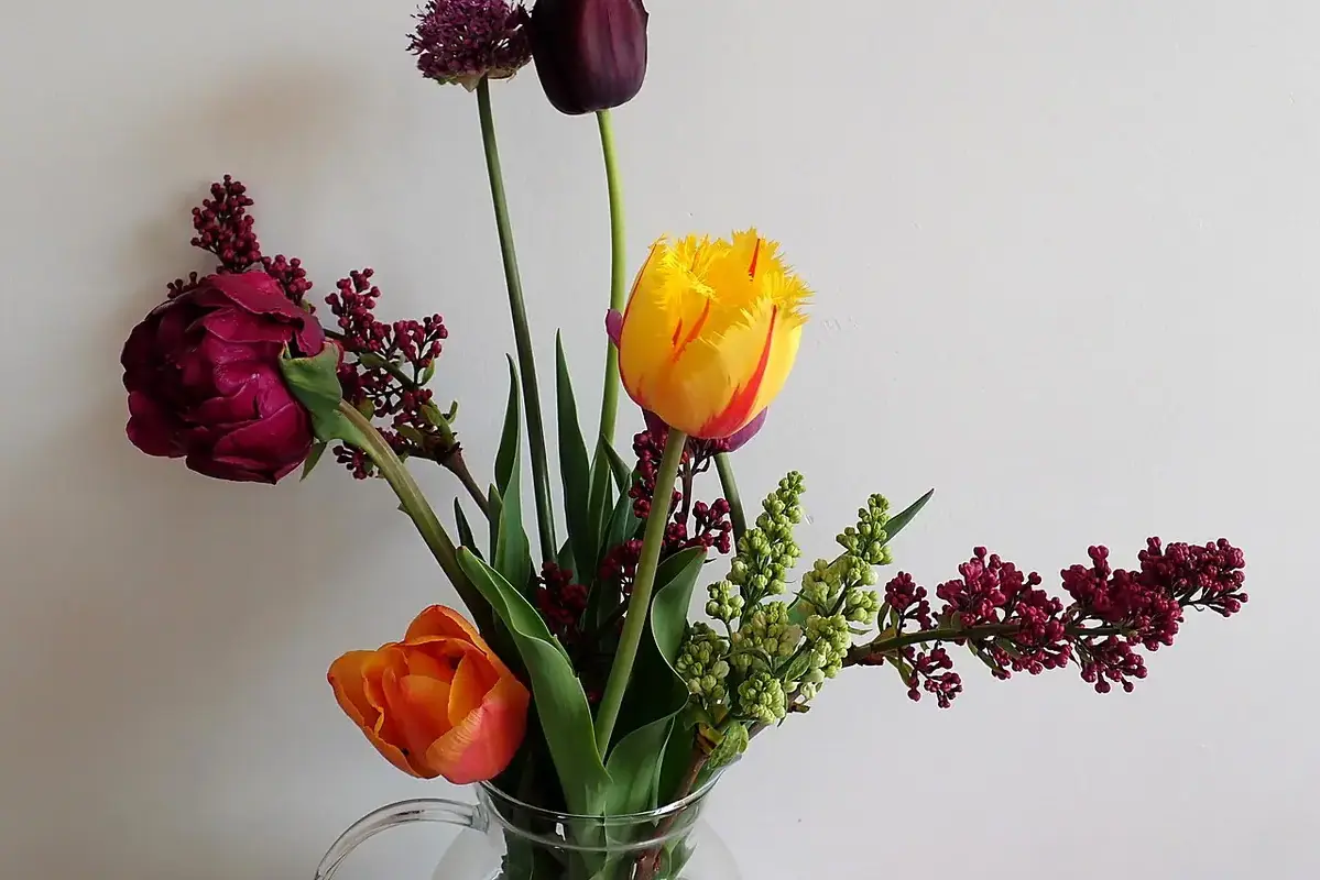Yellow and dark tulips arranged with lilac branches against a pale wall.